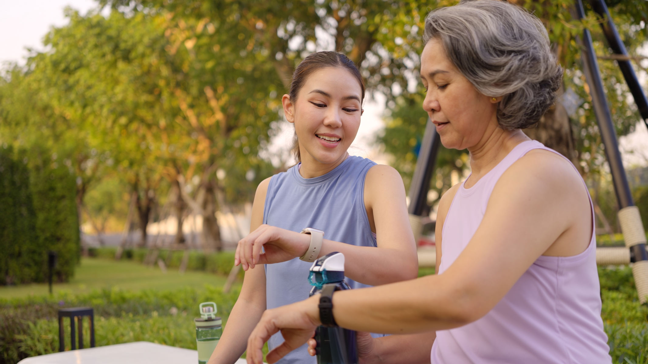 Two women are resting between exercises while sitting on a park bench outside and looking at their fitness trackers 