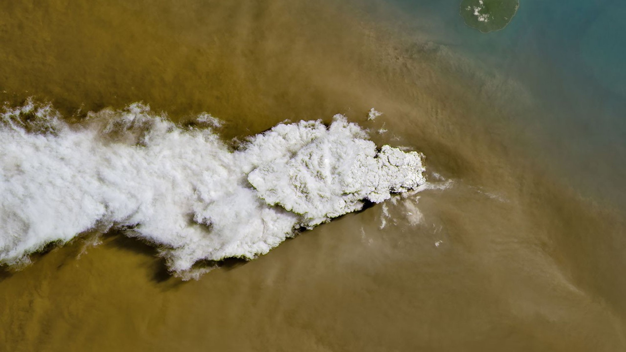 a plume of white-grey smoke rises from a small island, as seen from high overhead