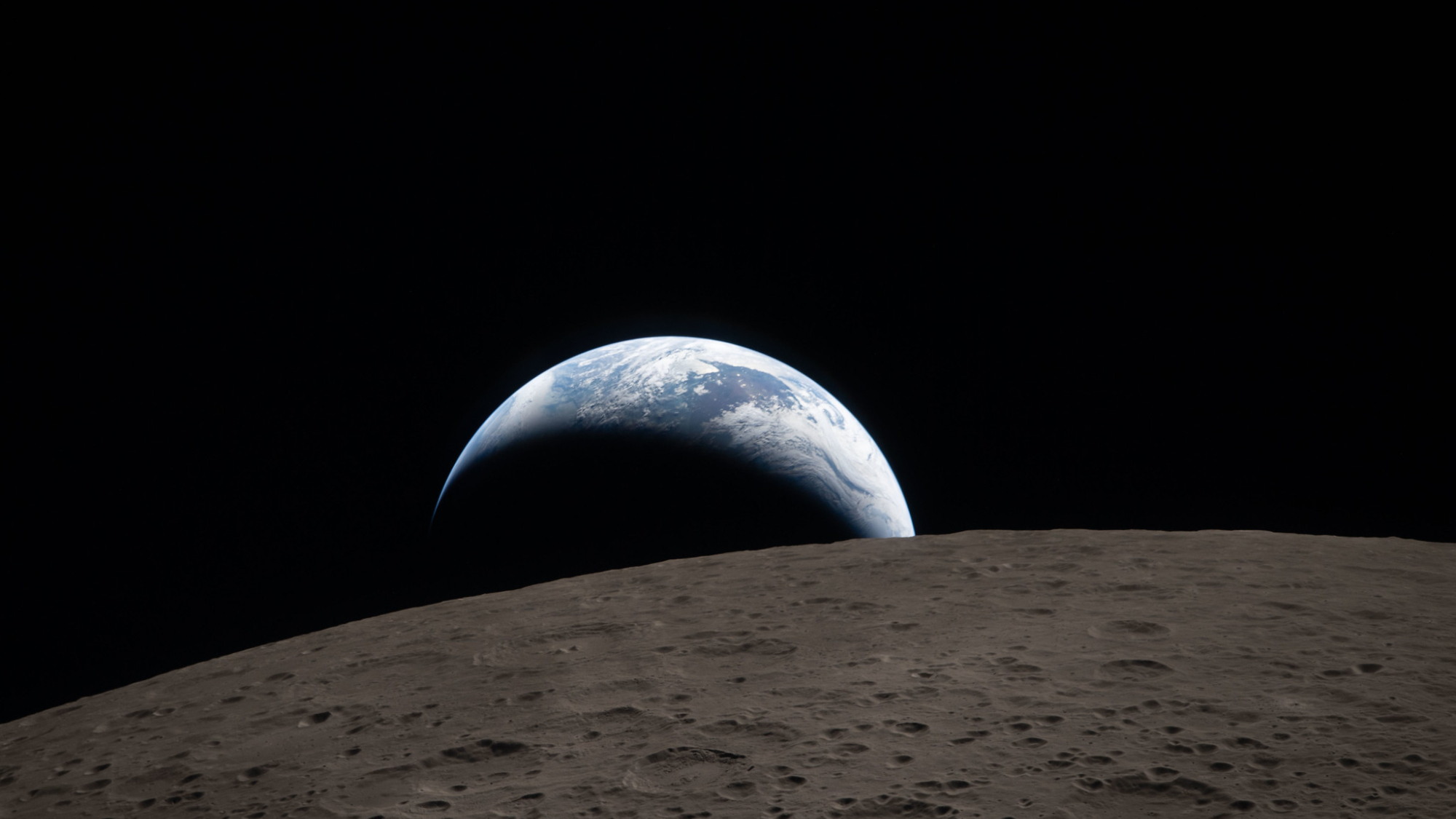 Earthset captured through the Orion spacecraft window at 6:41 p.m. EDT, April 6, 2026, during the Artemis II crew’s flyby of the Moon. A muted blue Earth with bright white clouds sets behind the cratered lunar surface.  The dark portion of Earth is experiencing nighttime. On Earth’s day side, swirling clouds are visible over the Australia and Oceania region.