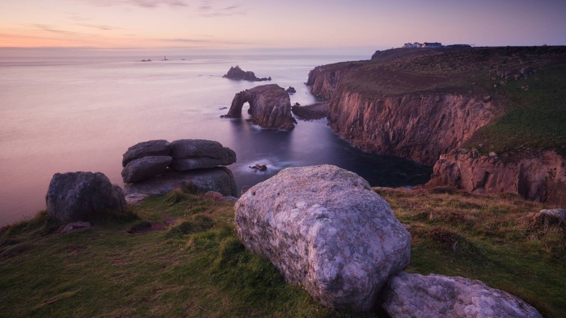 Rocky coastal cliffs (Land's End, Cornwall, UK) at sunset with a natural stone arch extending into the sea, grassy foreground, and calm water. In the far distance is a lighthouse.