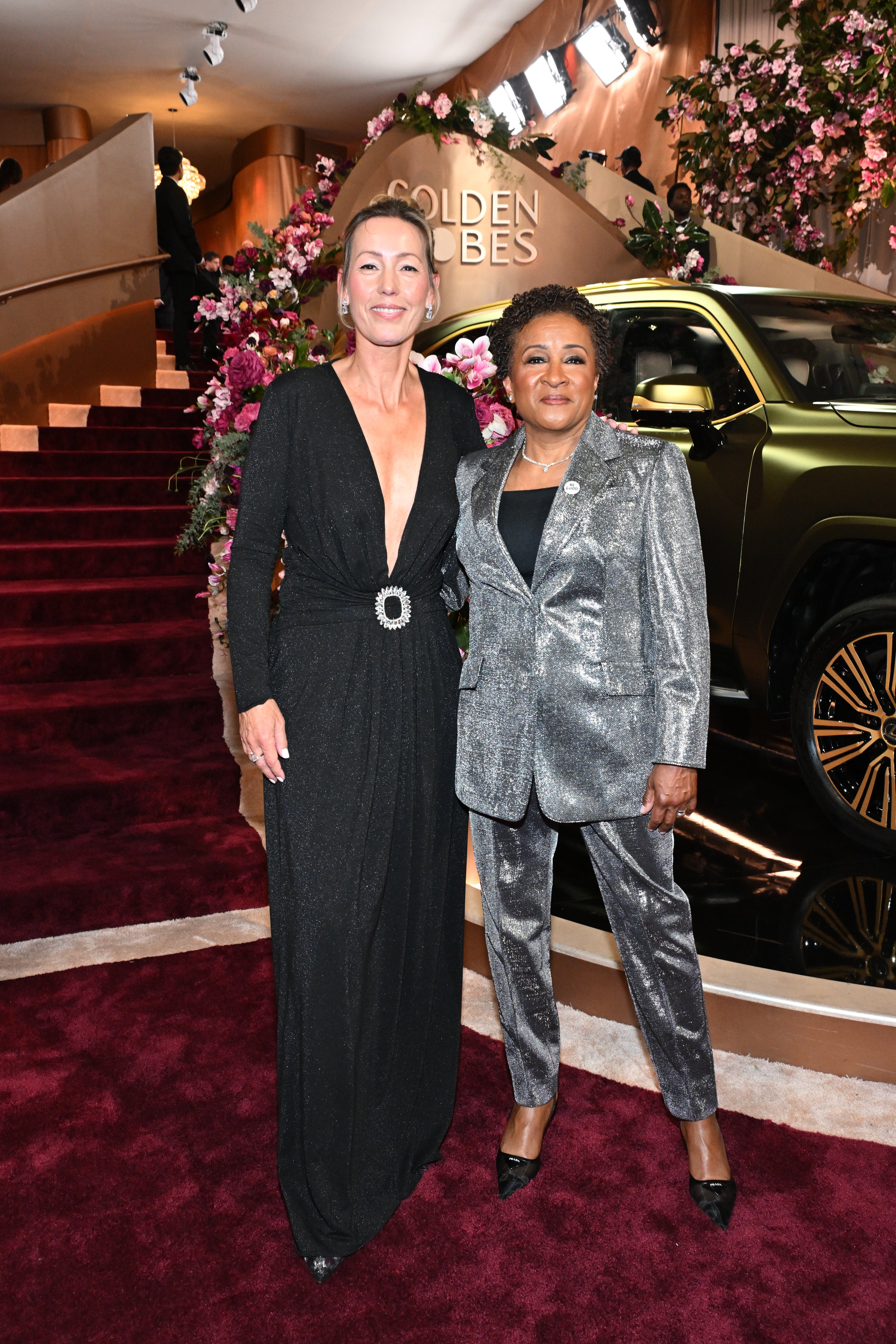 Two individuals on a red carpet. One wears a shimmering black gown, the other a silver suit. They stand in front of a "Golden Globes" sign