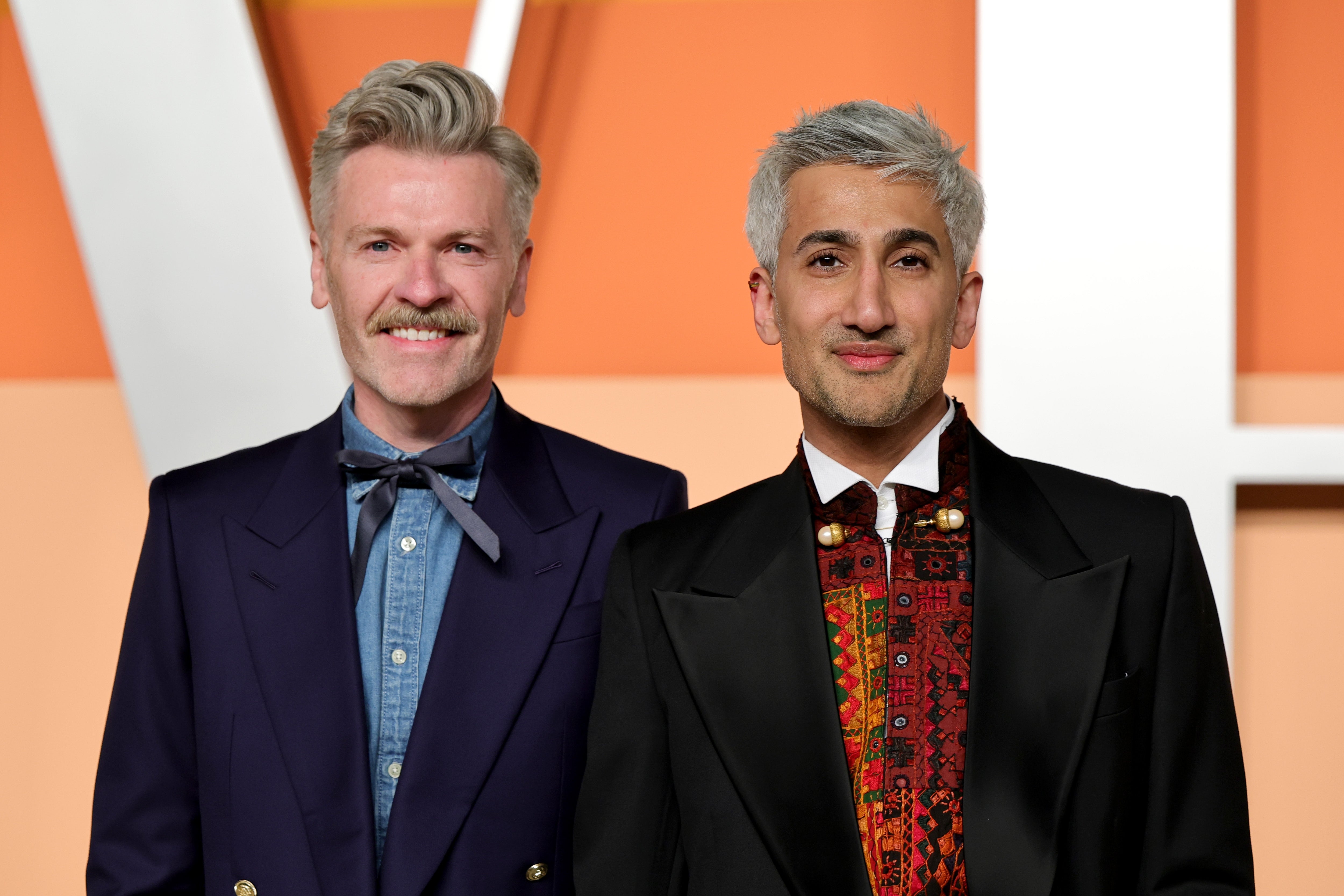 Two people at an event. One wears a suit with a denim shirt and bow tie, the other a tuxedo with a vibrant patterned shirt