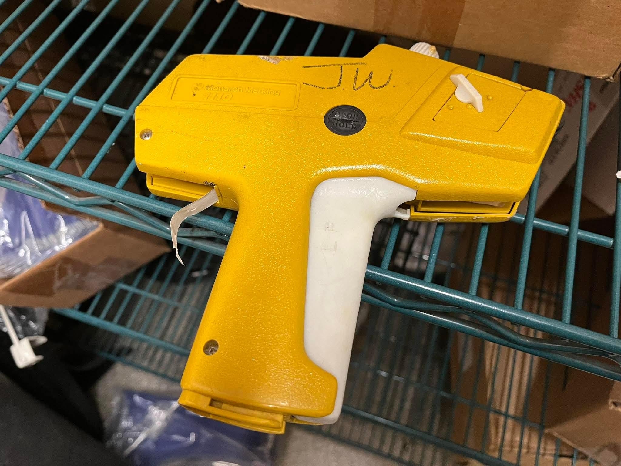 A yellow tagging gun with a white handle rests on a wire shelf